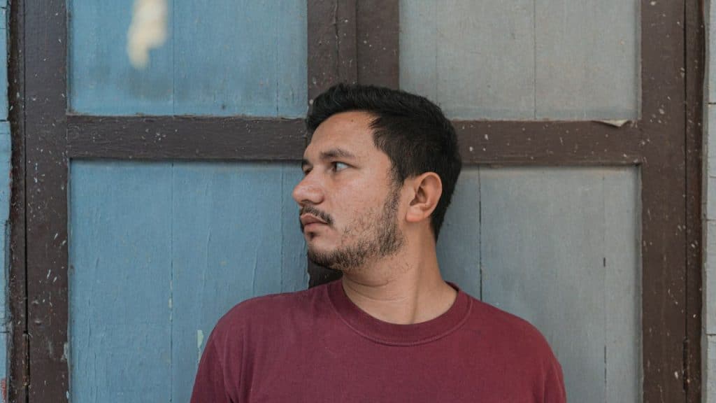 A man in a maroon shirt looks to his left against a blue and brown wooden wall.