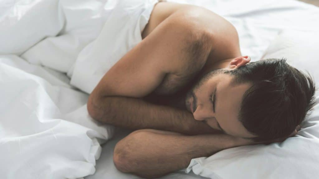 Man with bare torso sleeping on stomach, covered by white sheets.