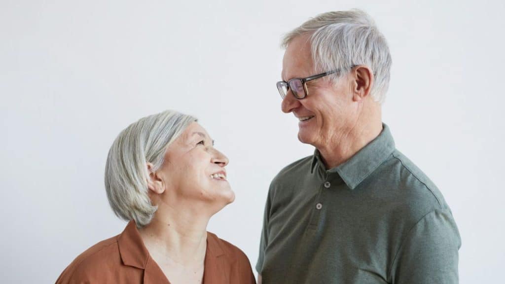 Two smiling elderly people with gray hair look warmly at each other.
