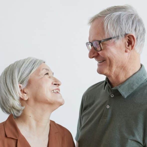 Two smiling elderly people with gray hair look warmly at each other.