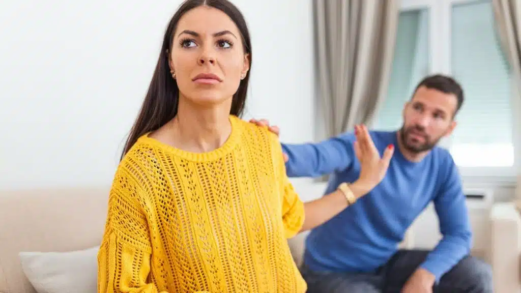Woman in a yellow top looks away as a man touches her shoulder and gestures.