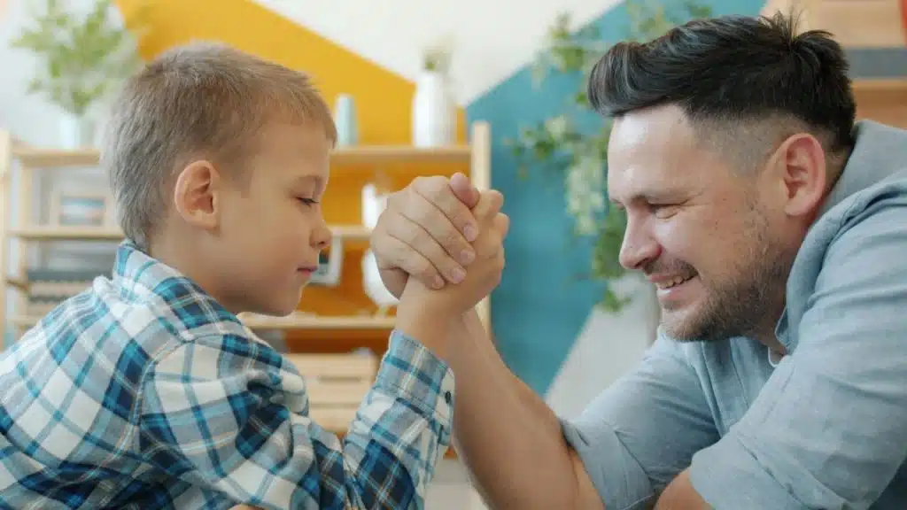 A man and a young boy, both smiling, engage in an arm-wrestling match in a brightly colored room.