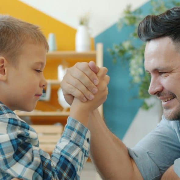 A man and a young boy, both smiling, engage in an arm-wrestling match in a brightly colored room.