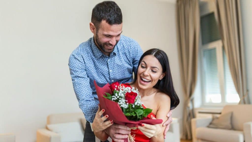 Smiling man giving a bouquet of red roses and baby's breath to a surprised and happy woman.