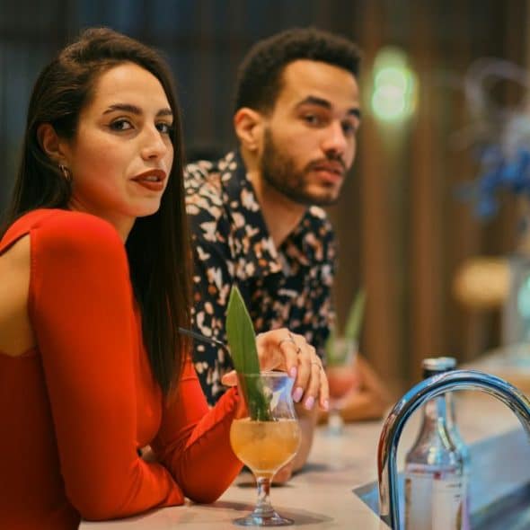 Woman in a red dress with a drink at a bar looking at the camera; man behind her.