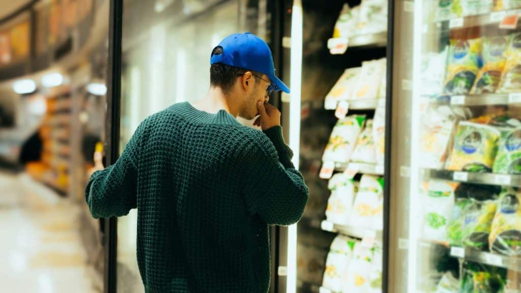 Man in a blue cap and green sweater looking into a supermarket freezer.