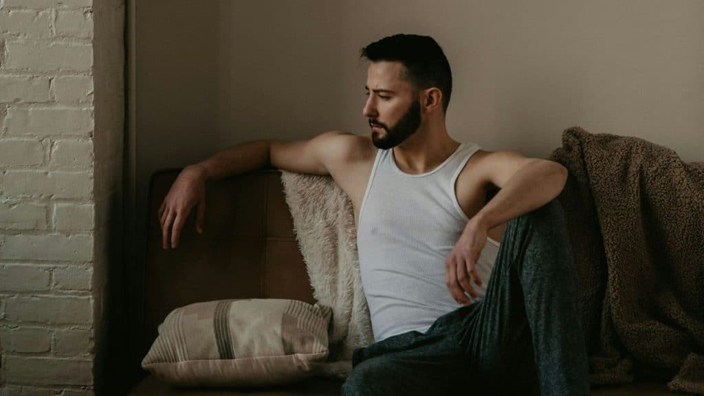 Bearded man in a white tank top sitting on a couch, looking away thoughtfully.