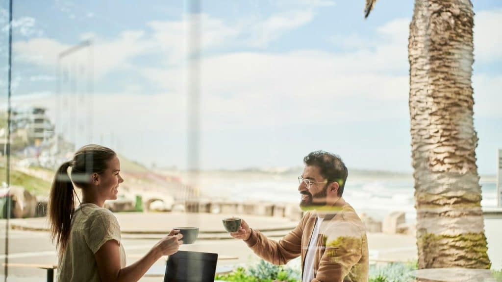 A man and woman smiling and having coffee at a beachside café.