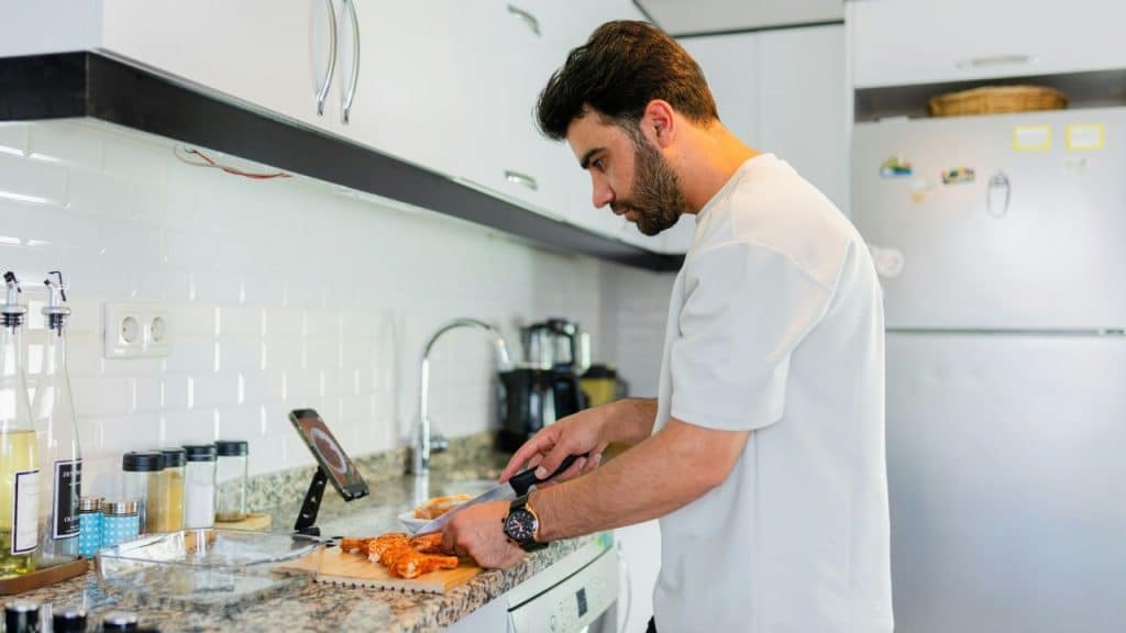 A man with a beard stands in a kitchen, looking at a smartphone on a stand while he cuts food on a cutting board.