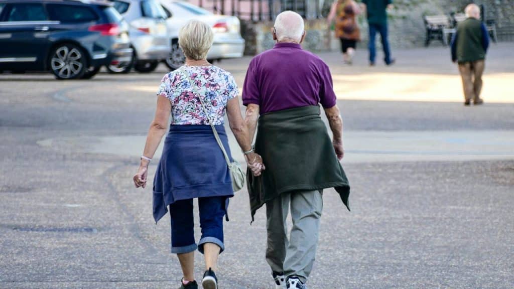 An older couple, viewed from behind, walks away while holding hands in a parking lot.