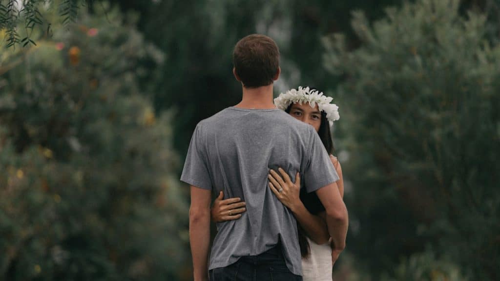 A couple embraces outdoors with the man's back to the camera, and the woman, wearing a white flower crown, looks over his shoulder.