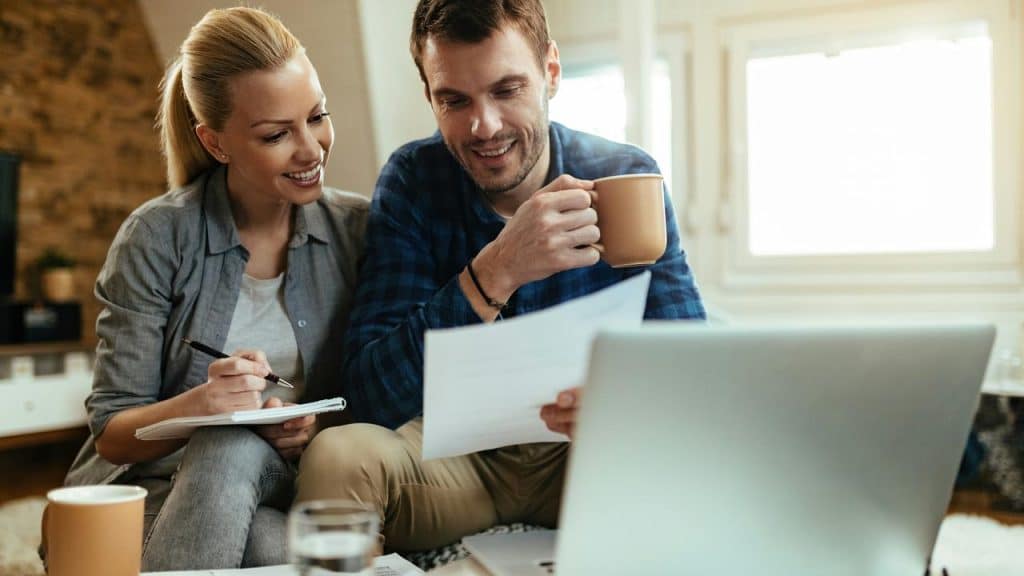 A smiling couple sitting together at a table reviewing documents with a laptop and coffee.