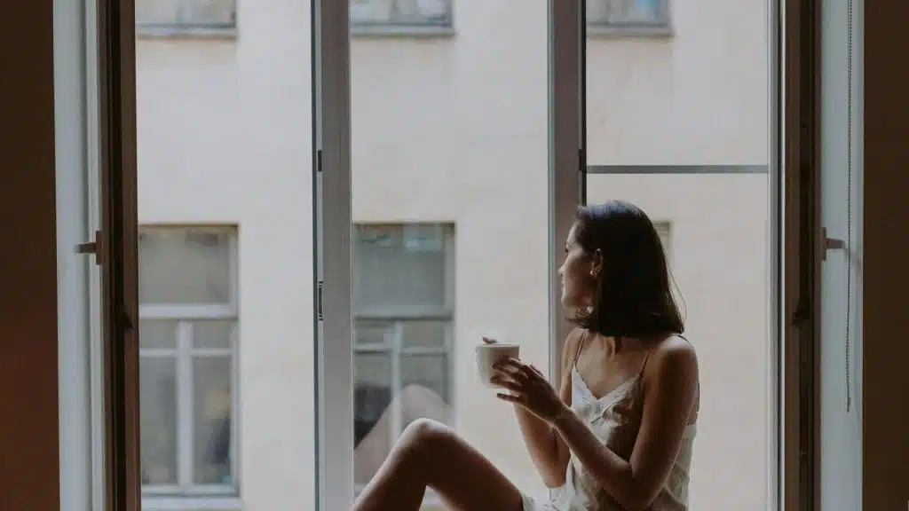 A woman sitting in a windowsill holding a cup and looking outside.