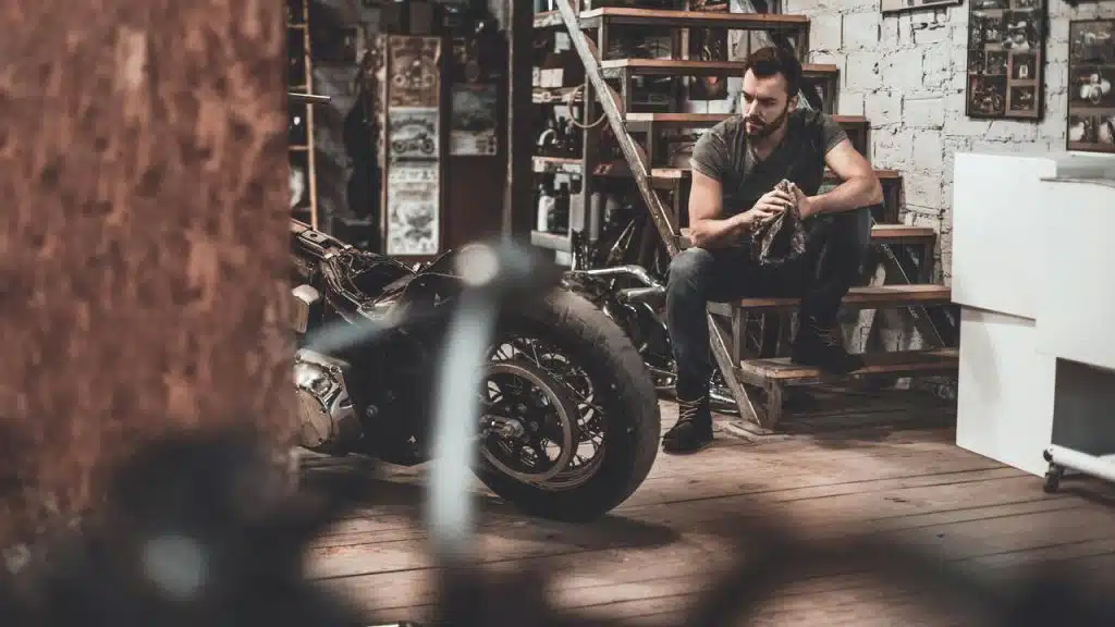 A man sitting in a workshop near a motorcycle, holding a cleaning cloth.