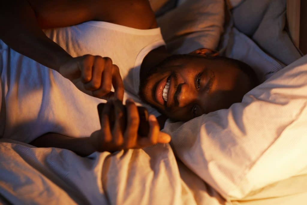 The man is smiling while using smartphone in bed.