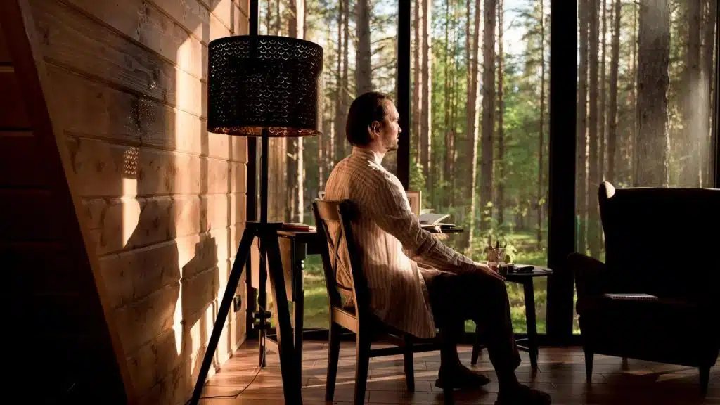 A person sitting by large windows in a cozy room looking outside.