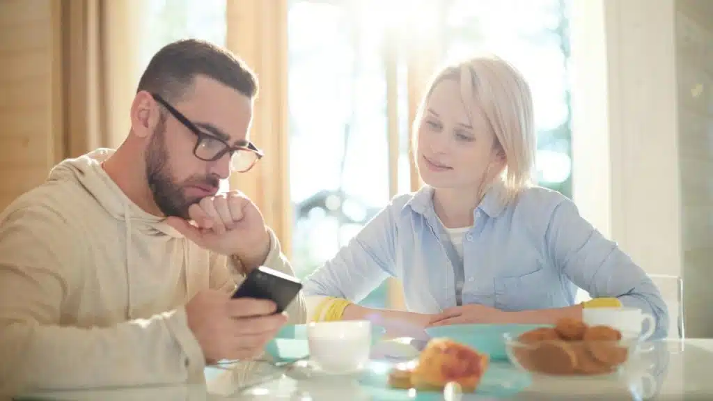 A man with glasses and a beard looks intently at his phone while a smiling blonde woman looks at him.