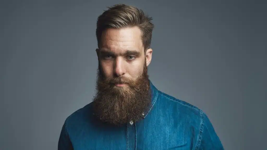A serious man with a full beard and denim shirt is looking down at the camera.