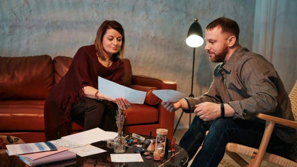 A man and woman are sitting and looking at papers across a coffee table with various items.