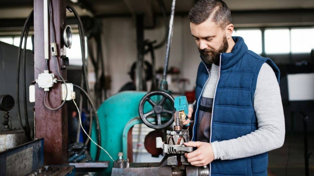 A bearded man in a blue vest is working on a machine part in a workshop.