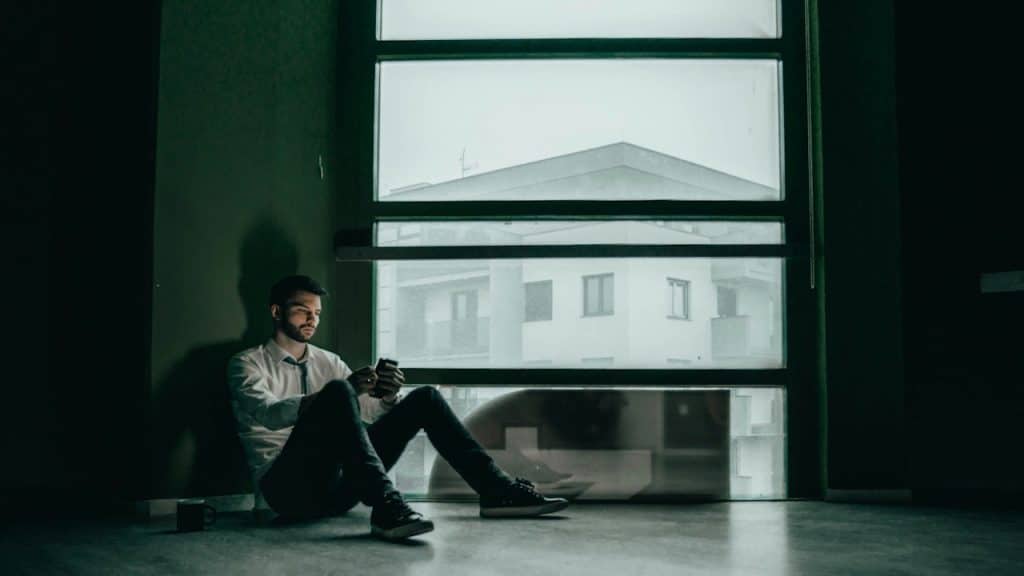 A solitary man in a white shirt and tie sits on the floor in a dark room, looking at his smartphone next to a large window.