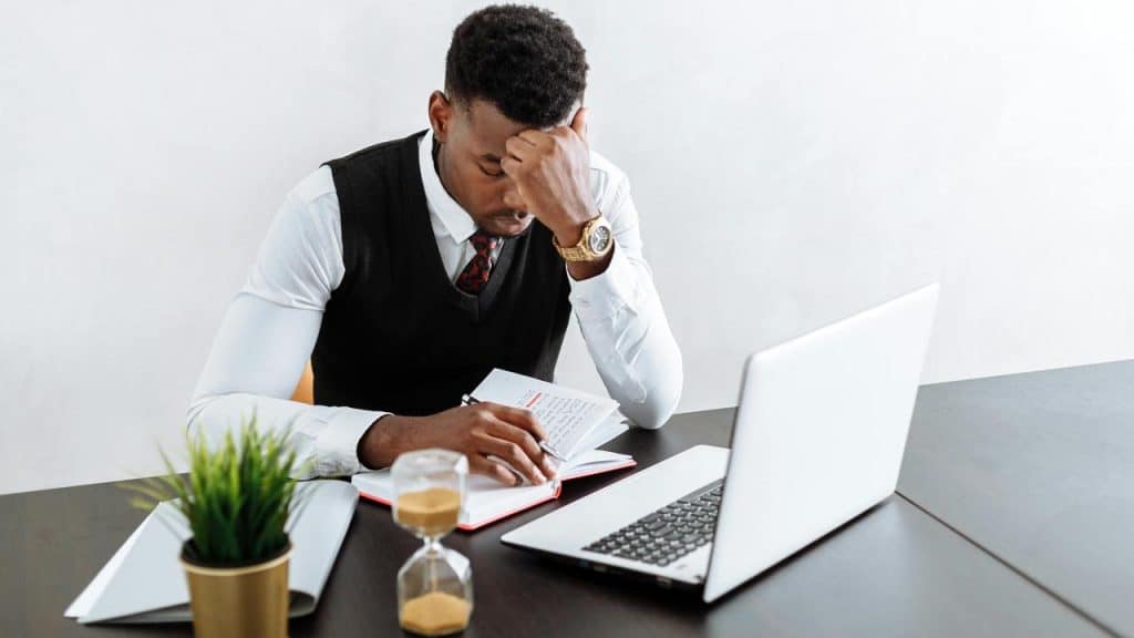 A man sitting at a desk with a laptop, looking stressed while writing in a notebook.