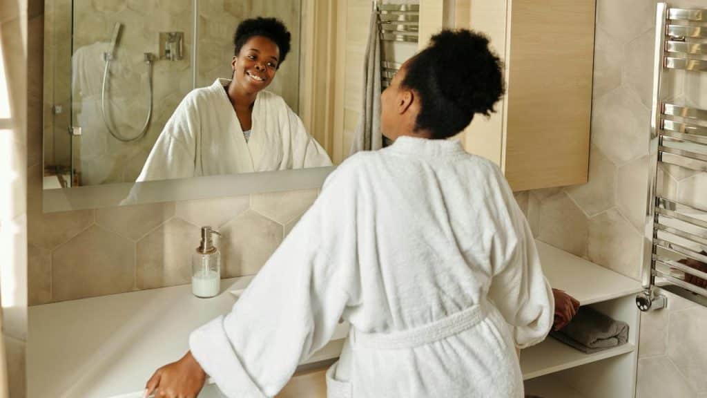 A woman in a bathrobe smiling at her reflection in a bathroom mirror.