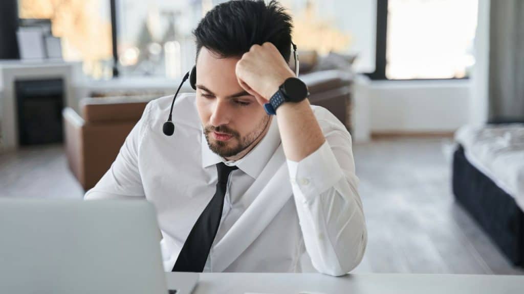A man in a shirt and tie, wearing a headset, looks tiredly at a laptop.