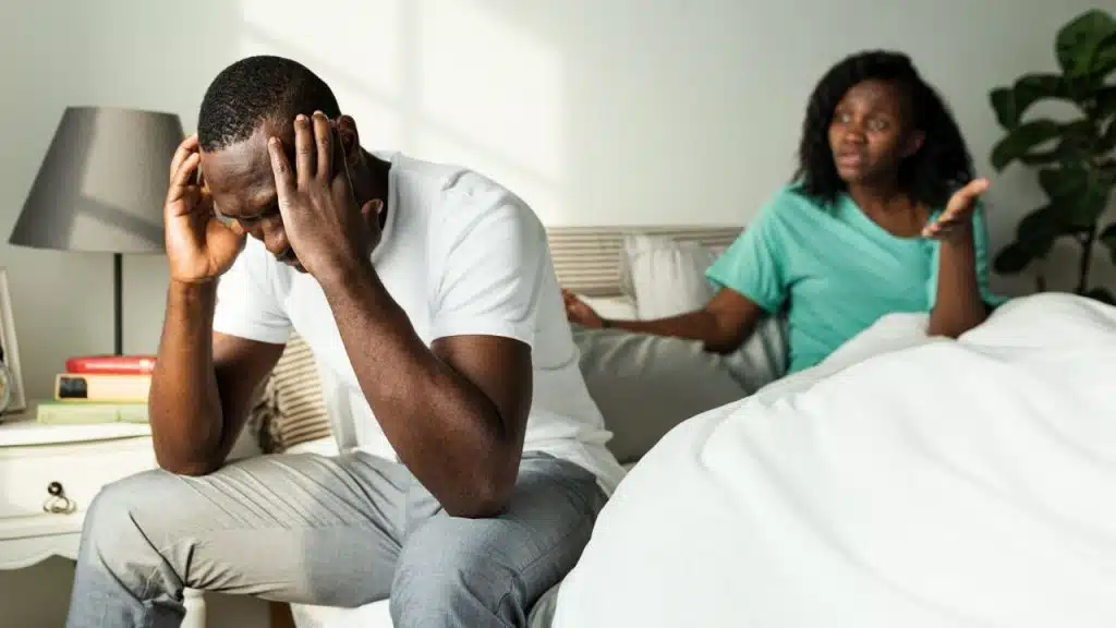 A Black man holds his head in distress as a Black woman argues with him in the background.