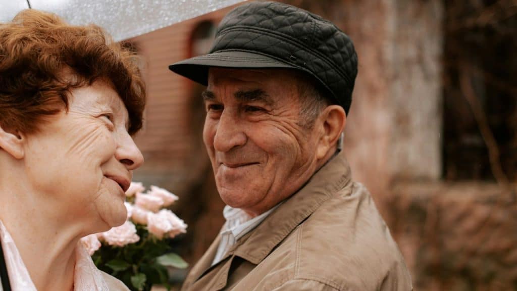 An elderly man in a cap smiles at an elderly woman holding pink roses outdoors.