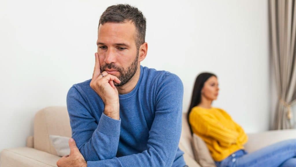 A worried man in a blue sweater sits with his hand on his cheek, facing away from a blurred woman.