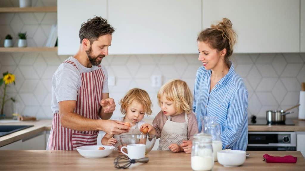 A couple and two small children in aprons are cracking eggs in a modern kitchen.