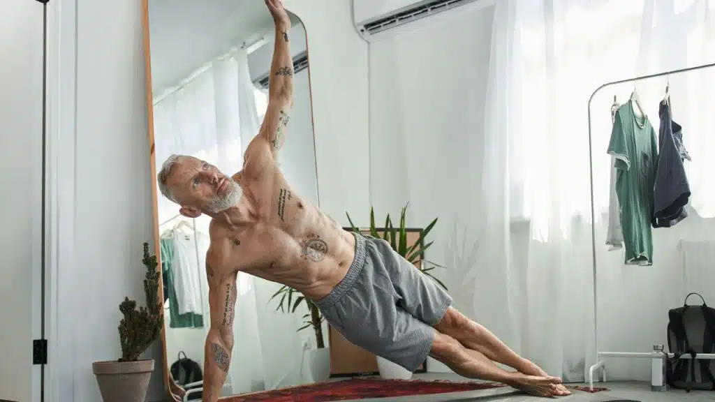 A fit, gray-haired man with tattoos is doing a side plank in a bright room.