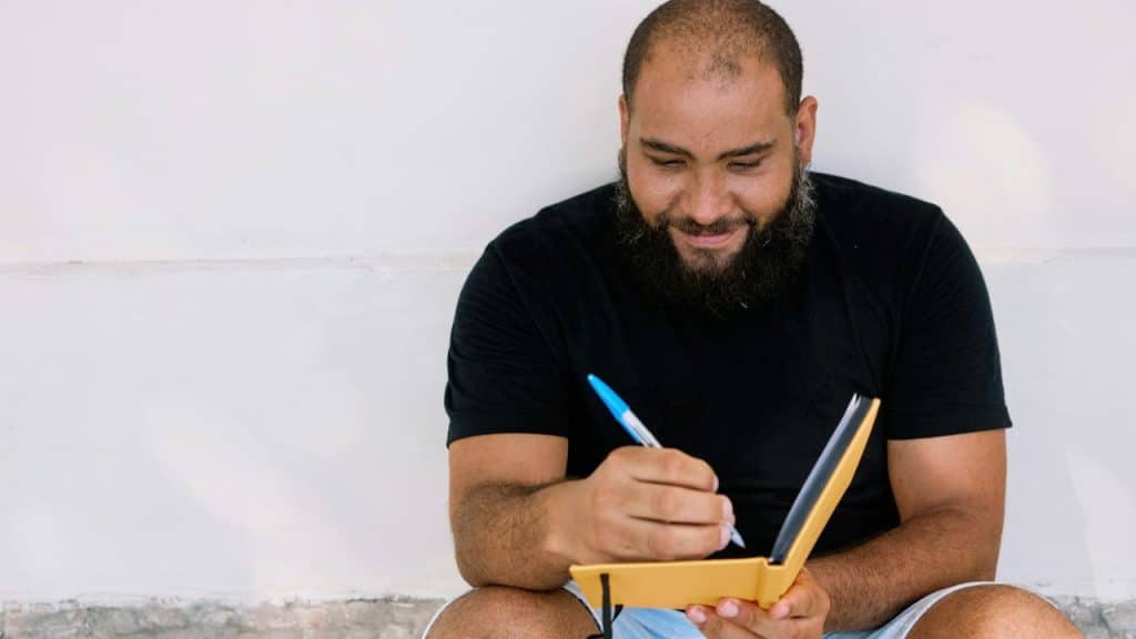 Smiling man with a beard and shaved head, wearing a black t-shirt, writing in a yellow notebook.