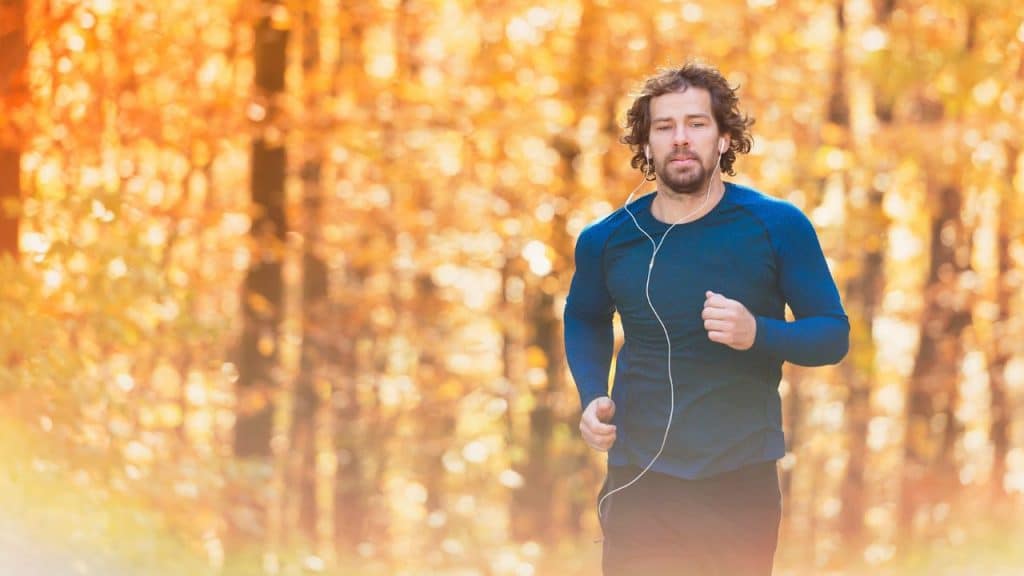 A man with earbuds runs through a sun-drenched forest with autumn orange foliage.