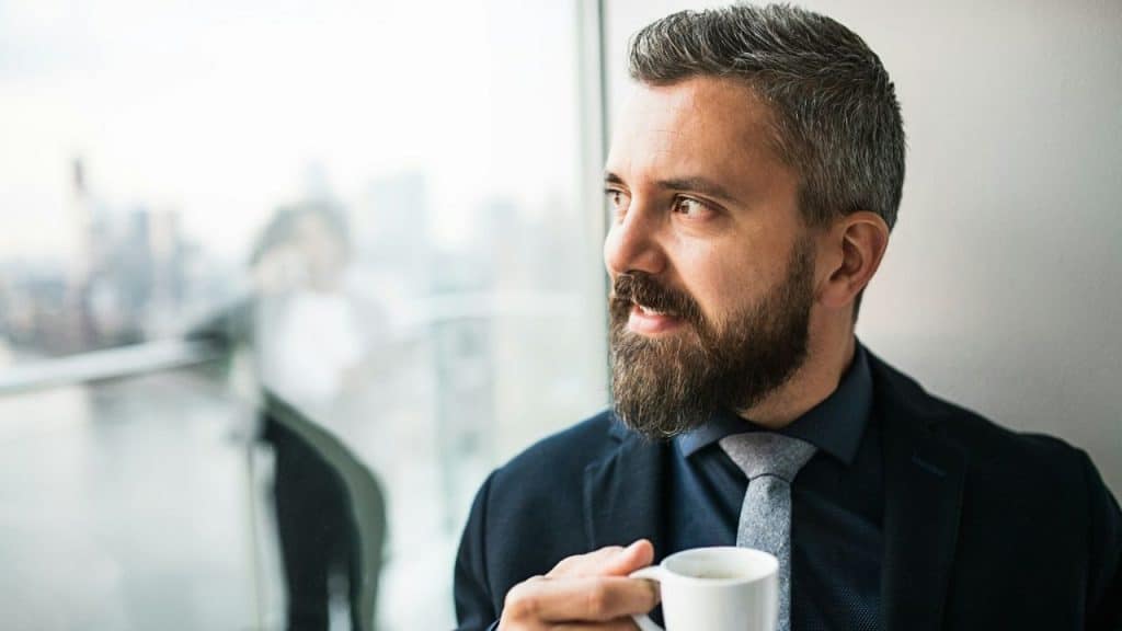 Man with beard in a suit holding a cup, looking out a window at a city.