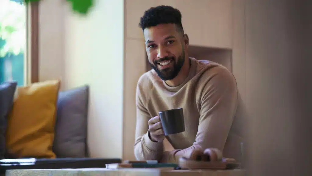 Smiling man with a beard holds a dark mug while sitting at a table near a window.