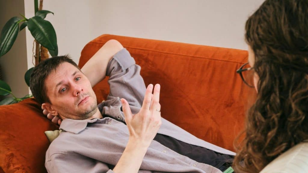 A man is reclining on an orange couch, talking and gesturing to an unseen person.