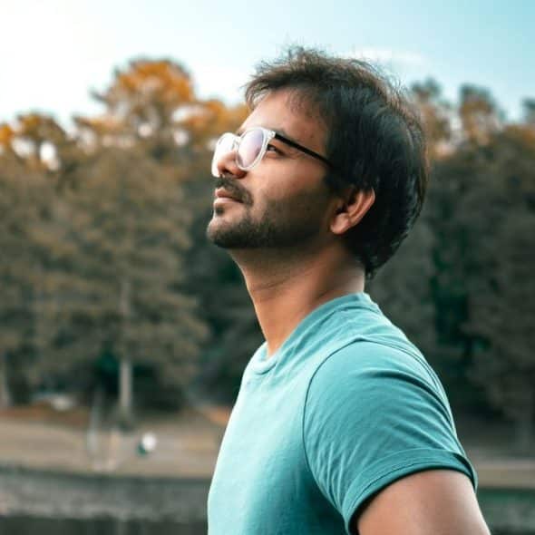 Man in teal shirt and glasses looking up against a blurry outdoor background.
