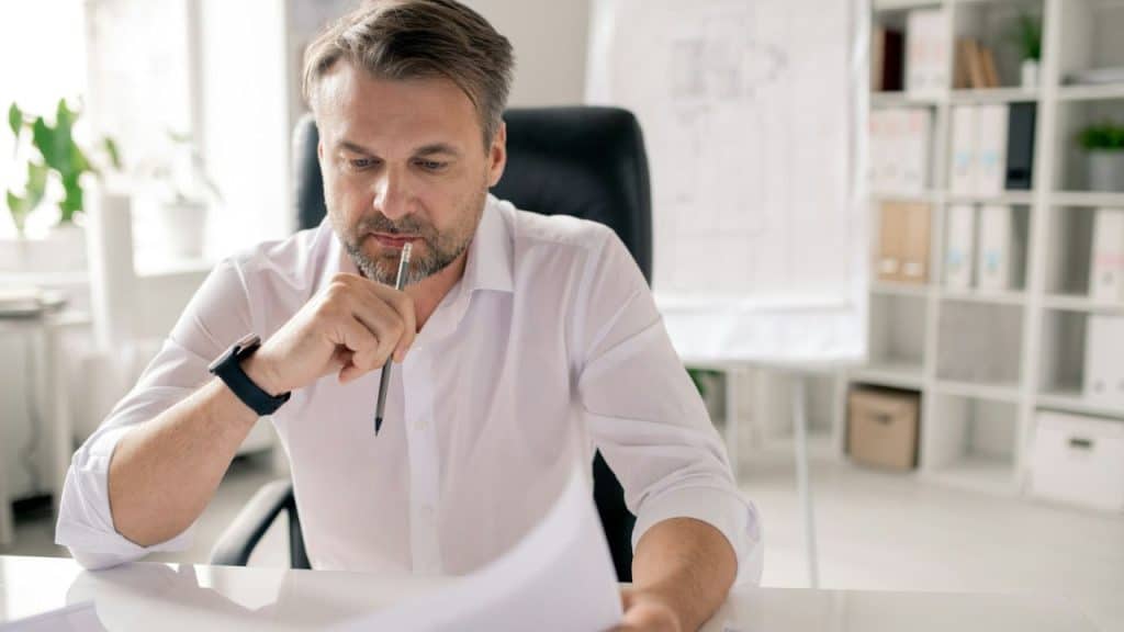 Middle-aged man in a white shirt sitting at a desk, looking down at papers, holding a pencil to his chin.