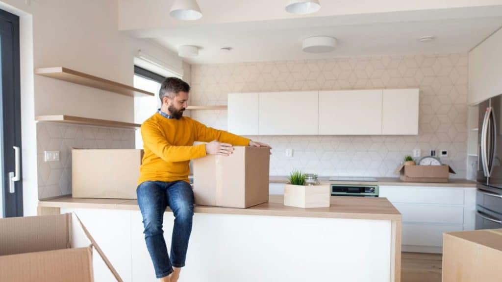 A man sits on a kitchen counter, unpacking a cardboard box in a bright, modern room.