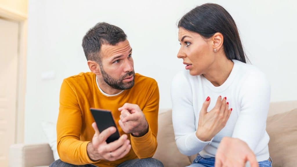 A man holds a phone and talks to a woman who is gesturing with her hands.