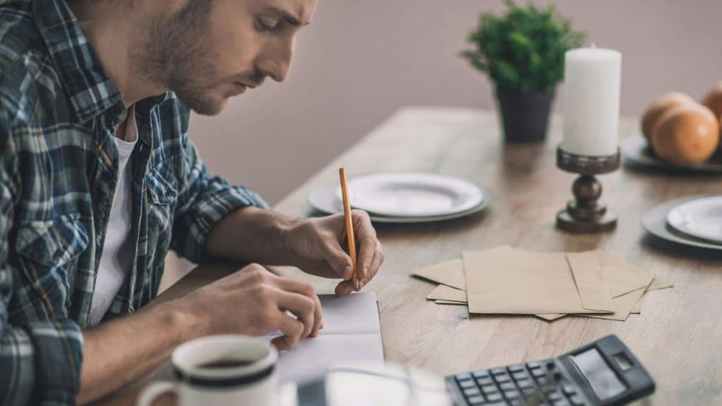 A man in a plaid shirt sits at a table writing with a pencil and calculator nearby.