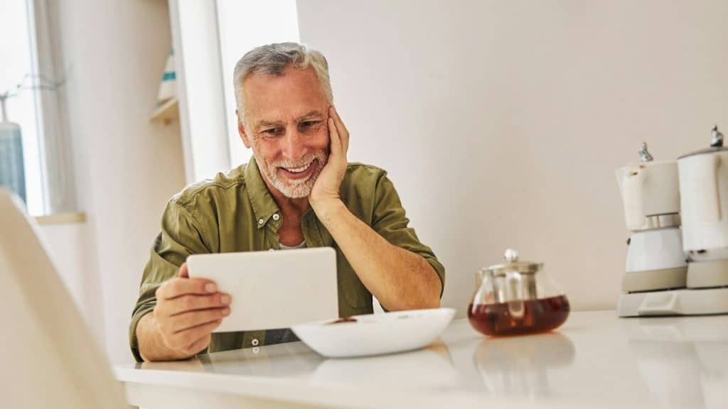 Smiling senior man holding a tablet with his hand on his cheek at a kitchen table.