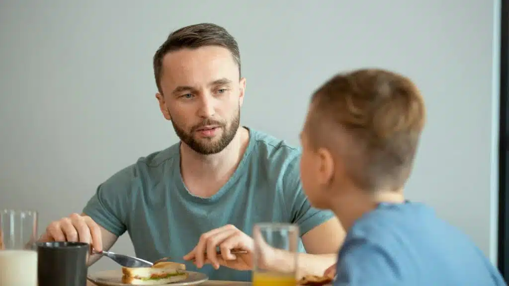 Man in a teal shirt having a meal, talking to a boy who is out of focus.