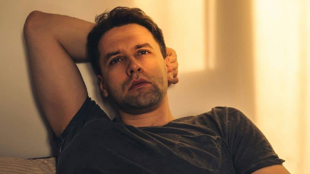 A serious man in a dark t-shirt is resting with his arm behind his head, looking forward.