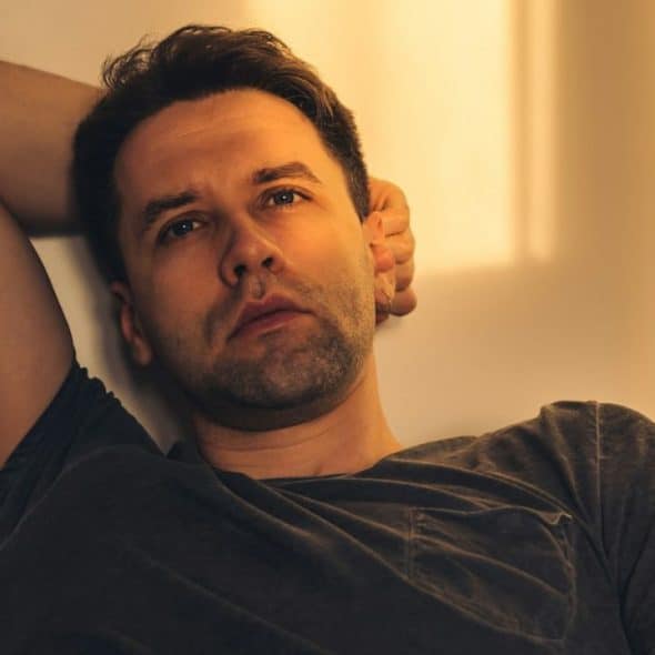 A serious man in a dark t-shirt is resting with his arm behind his head, looking forward.