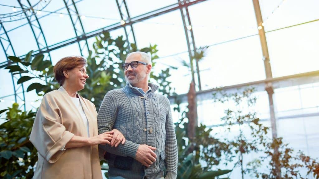 An older man in a gray sweater and a woman in a light jacket walking arm-in-arm through a sunny, tropical-like indoor garden.