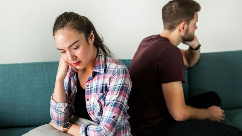 A woman in a plaid shirt sits on a teal couch, looking down unhappily, with a man sitting next to her with his back turned.