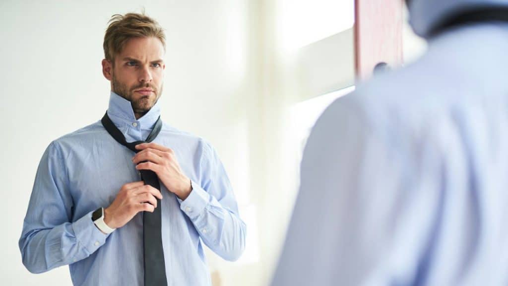 A focused man with a beard is tying a dark tie in front of a mirror while wearing a light blue collared shirt.